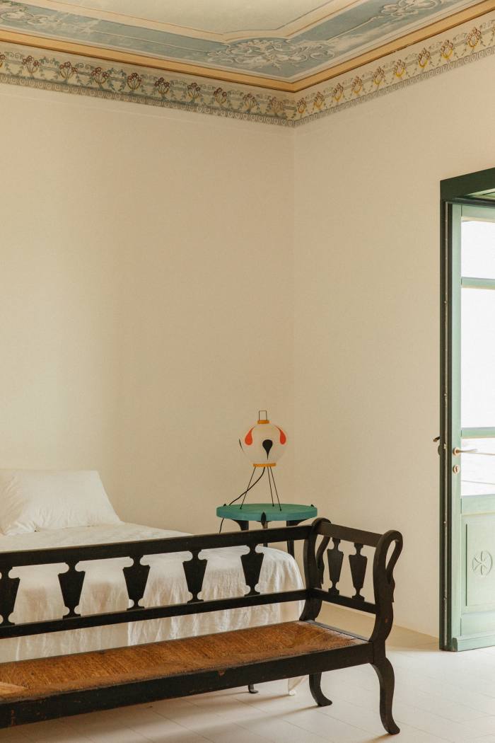 Una panca in rattan nero ai piedi del letto, trovata da Kapoor in un mercato in Italia.  Il comodino è di Ashley Hicks e la lampada di Noguchi