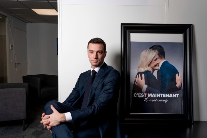 Jordan Bardella siede accanto a una grande foto incorniciata di lui che abbraccia Marine Le Pen