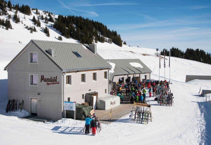 Ristorante Alprestaurant Panüöl circondato dalla neve, con una collina alle spalle, e tanta gente in tuta da sci seduta sulla terrazza