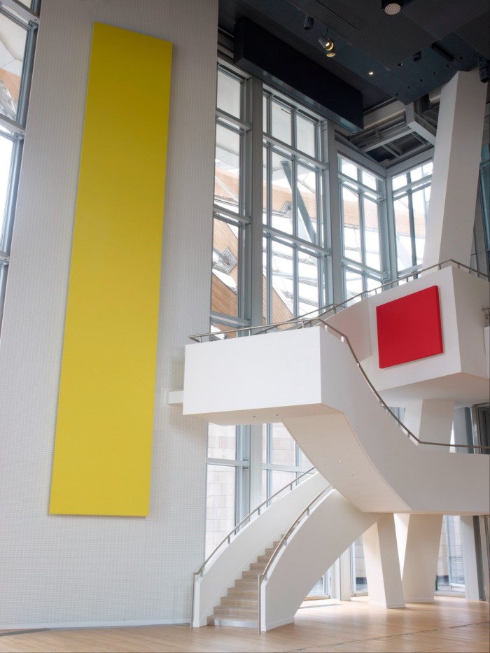 Una grande stanza con un soffitto alto, enormi finestre e una scala.  Le pareti sono dipinte di bianco con una grande striscia gialla su una e un quadrato rosso sull'altra