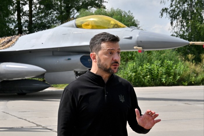 Volodymyr Zelenskyy gesticola mentre parla ai rappresentanti dei media mentre è in piedi di fronte a un aereo da caccia F16