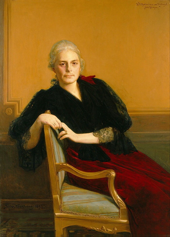 Una donna che indossa un abito rosso scuro bordato di pizzo nero è adagiata su una poltrona e guarda dritto verso l'artista