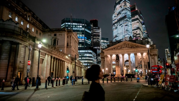 The image shows the City of London at night, featuring the illuminated Royal Exchange and the Bank of England