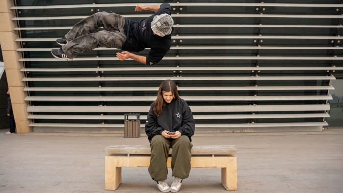 A young man performs a backflip over a woman sitting on a bench, absorbed in her phone, in Tehran