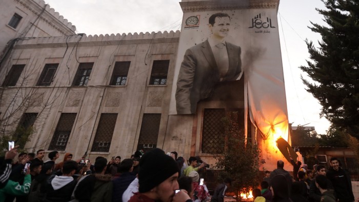 Flames can be seen licking up from the bottom of a poster featuring a large image of a smiling Bashar al-Assad on the side of a building in Hama on Thursday