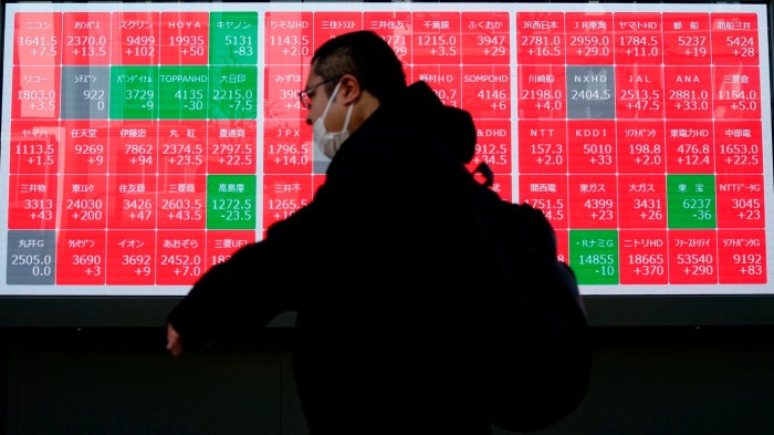 A person wearing a mask walks past an electronic stock board displaying the Nikkei index