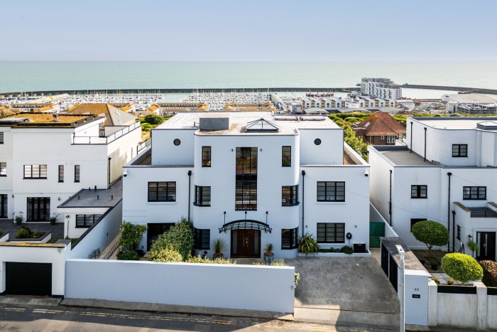 Una casa bianca in stile Art Déco con vetrate curve, un atrio centrale in vetro e una tettoia d'ingresso con cornice nera, situata vicino a un porto turistico con file di barche e affacciato sul mare, fiancheggiata da proprietà moderne simili e giardini paesaggistici