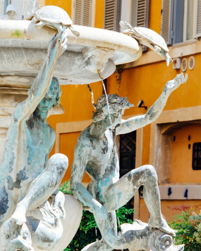 Dettaglio di Fontana Delle Tartaughe: due statue maschili che si allungano verso le tartarughe di pietra appoggiate sul bordo di un bacino della fontana, con un edificio residenziale arancione sullo sfondo