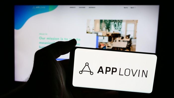 A person is holding a smartphone with the logo of AppLovin in front of website