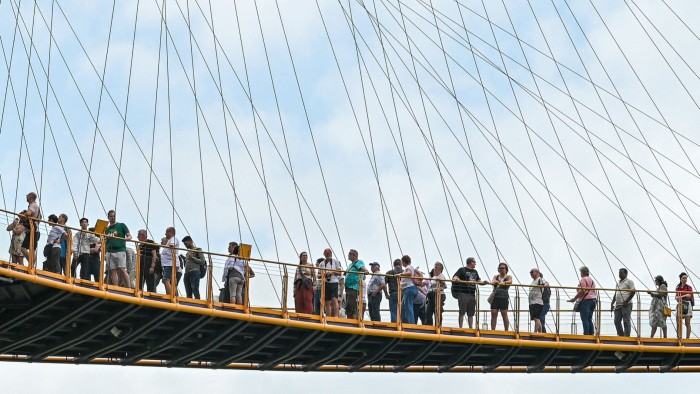 A walkway suspended high above the ground and supported by numerous cables is bustling with people enjoying the view against a clear sky