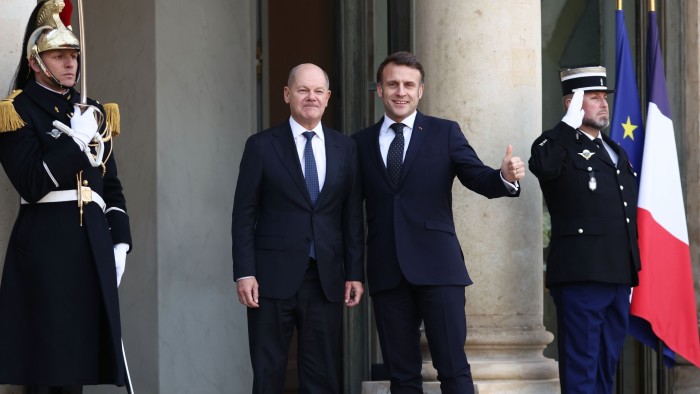 Emmanuel Macron and Olaf Scholz stand side by side, smiling, in front of the Élysée Palace in Paris.