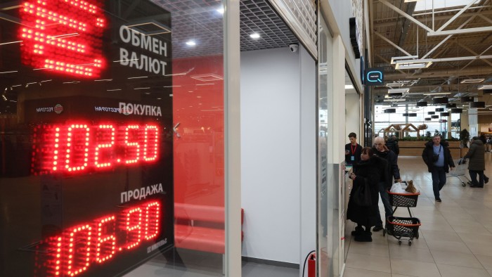 People walk past an exchange office with an electronic panel showing currency exchange rates against the rouble