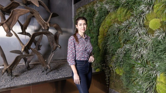 A woman in a floral shirt and jeans stands next to a green plant wall and a metal bird sculpture in an indoor setting