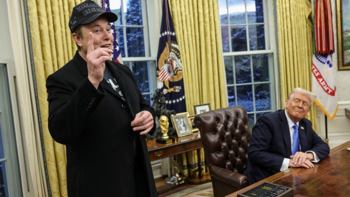 Elon Musk, wearing a ‘Make America Great Again’ cap, speaks while standing in the Oval Office next to Donald Trump, who is seated at the Resolute Desk, smiling and listening