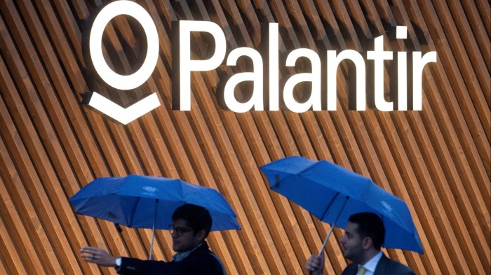 Two men holding blue umbrellas walk past the illuminated sign of Palantir Technologies on a wall