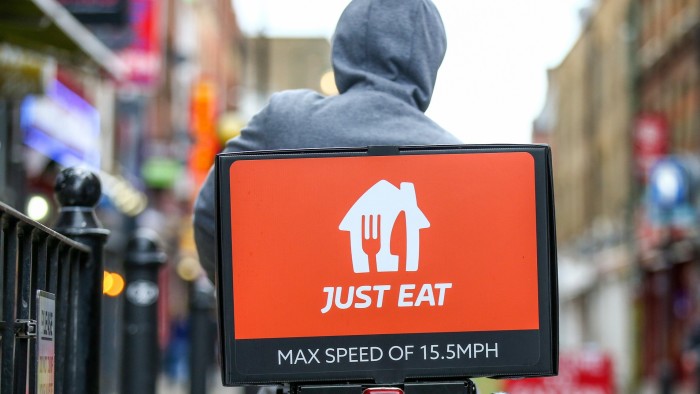 Back view of a food delivery courier wearing a grey hoodie riding a bicycle on Brick Lane in London. The delivery box on the back is branded with the "Just Eat" logo in white on an orange background, and it includes the text "MAX SPEED OF 15.5 MPH."