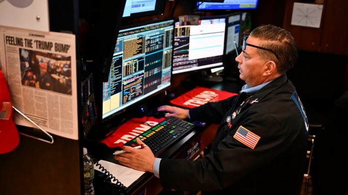 The trader has “Trump” mats, stickers and a newspaper cutting around his workstation. His glasses are perched on his forehead