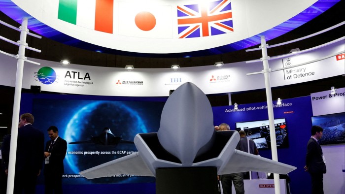 A fighter jet displayed prominently at a defence show, with the model positioned in front of a booth featuring logos of participating companies and organisations, including the Acquisition, Technology & Logistics Agency, Mitsubishi Heavy Industries, and the Ministry of Defence. National flags of Italy, Japan, and the UK are visible above the display.