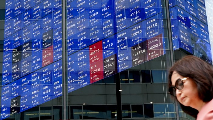 A woman wearing sunglasses walks past an electronic board displaying the Nikkei 225 index with various stock prices and values