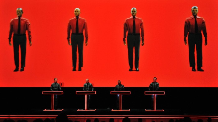 Four men stand at lecterns on a stage. Behind them, large images of the four men, dressed identically in suit trousers, shirts and ties, are shown on a bright red screen