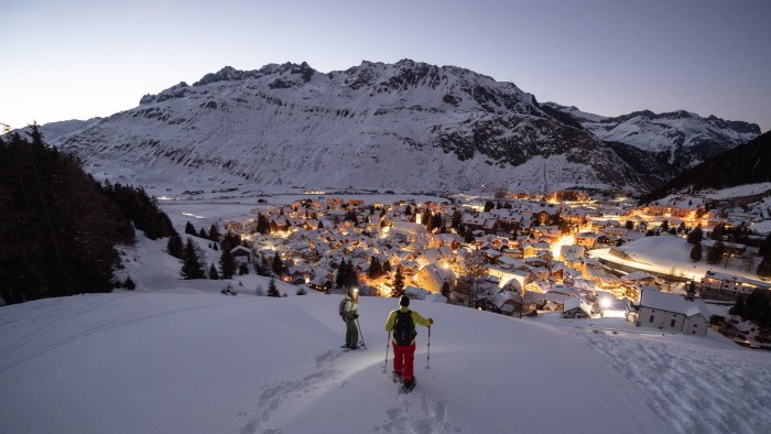 Two people are snowshoeing on a snowy hill overlooking Andermatt at night