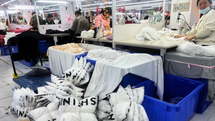 Bags branded “Shein” lie bundled together on the floor in front of where the women are working