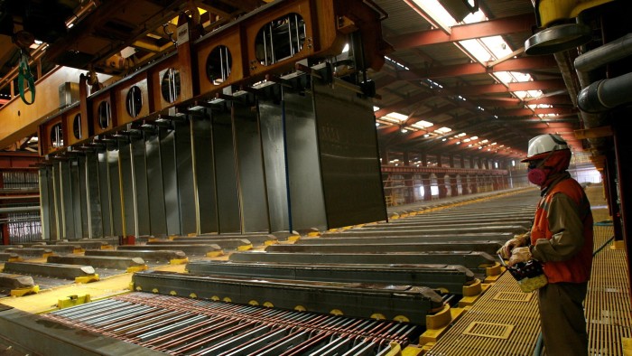 A worker at BHP's Escondida copper mine in Antofagasta, Chile, stands inside a copper cathodes plant with large metal sheets suspended from machinery and the worker wearing protective gear, including a helmet and mask