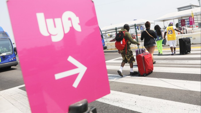 Travelers with luggage walk towards a Lyft pickup area at Los Angeles International Airport. A prominent pink Lyft sign with an arrow directs them.