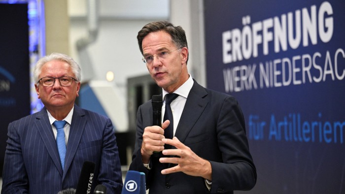Mark Rutte speaks holding a microphone next to Armin Papperger at the inauguration of Rheinmetall’s new artillery plant.