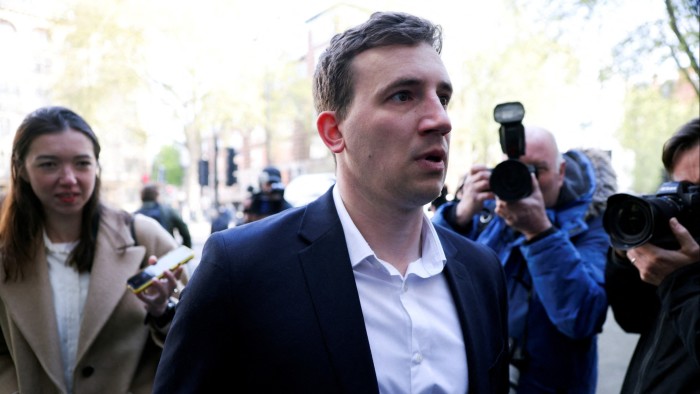 Christopher Cash arrives at Westminster Magistrates' Court, surrounded by photographers and a reporter holding a phone.