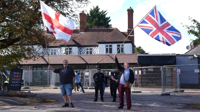 Two protesters hold large English and Union Jack flags outside the Bell Hotel, with several police officers standing behind them.