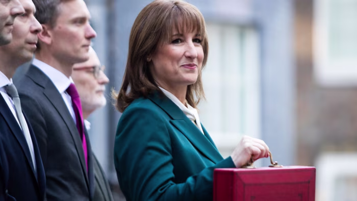 Rachel Reeves stands outside holding the red Budget box, with several suited colleagues beside her