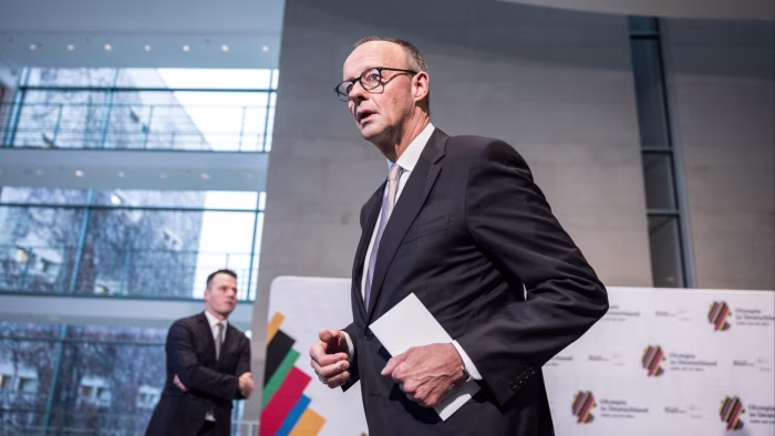 Friedrich Merz holds a folder and walks away after signing an agreement, with another man standing in the background.