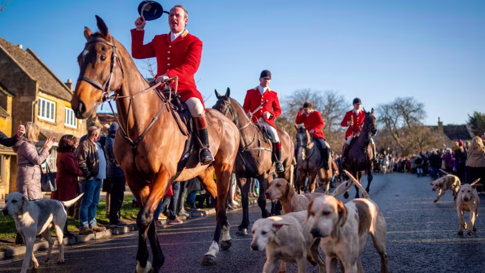 Horse riders in red jackets and packs of hounds parade down a street lined with spectators during the North Cotswolds Hunt.