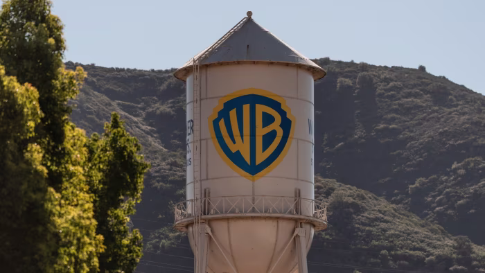 The Warner Bros water tower with the WB shield logo stands in front of green hills and trees.