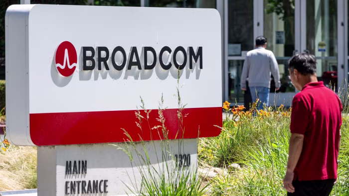 Broadcom headquarters sign with logo and text, with two people walking nearby and office doors in the background.