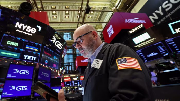 A trader in a jacket with a U.S. flag patch works on the NYSE floor, surrounded by digital stock ticker screens.