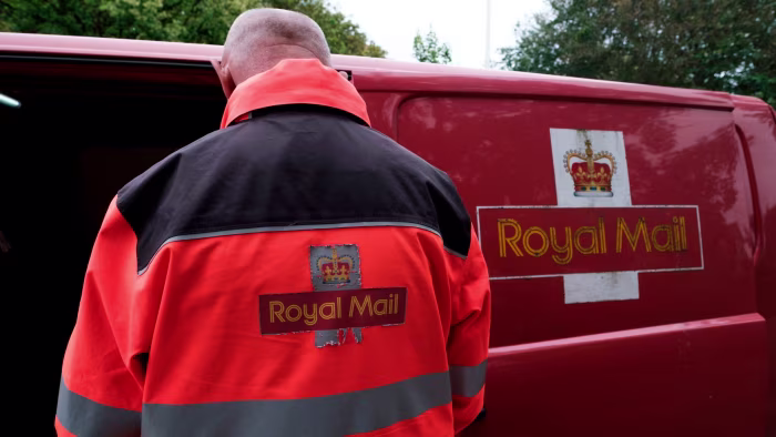 A Royal Mail employee in a red and black uniform sorts mail at the back of a delivery van displaying the Royal Mail logo.