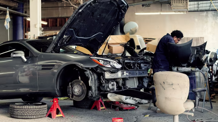 A mechanic repairs a car with its hood open and front bumper removed at an auto shop, surrounded by tools and car parts.