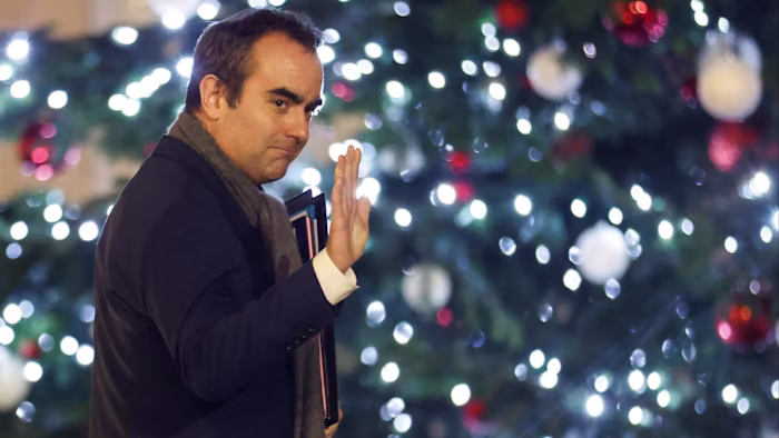 Sébastien Lecornu waves while leaving the Élysée Palace, holding documents, with a decorated Christmas tree in the background.