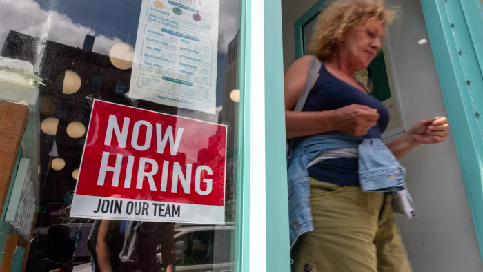 A "Now Hiring" sign is displayed in a business window as a woman exits the storefront.