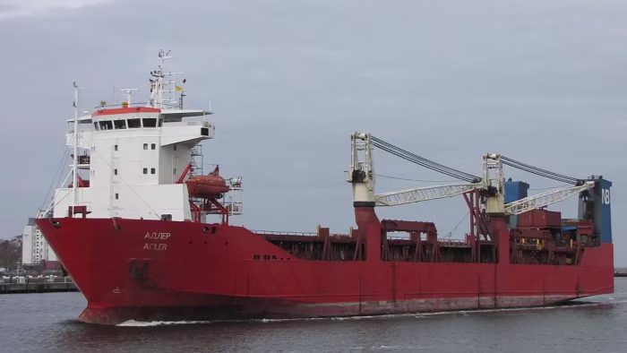 The Russian cargo ship Adler sails on the water, showing a red hull, white superstructure, and onboard cranes.