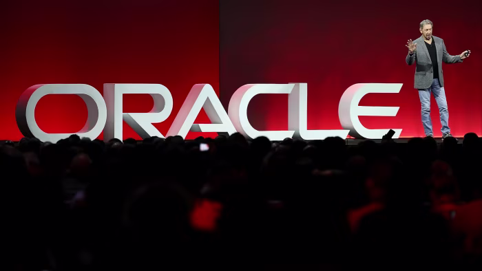 Larry Ellison stands on stage in front of a large ‘ORACLE’ sign, delivering a keynote to an audience