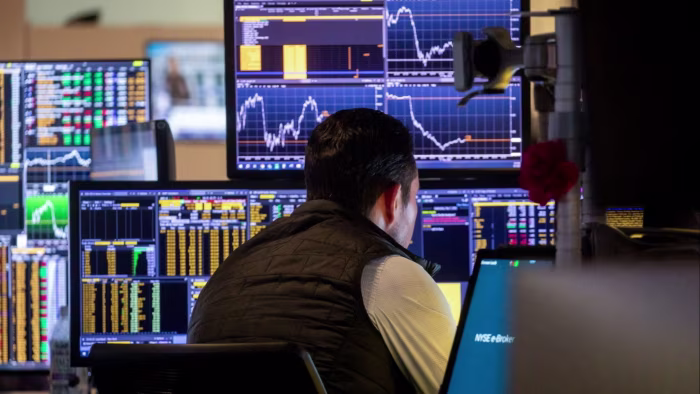 A trader sits at a desk surrounded by multiple monitors displaying stock charts and financial data on the NYSE trading floor.