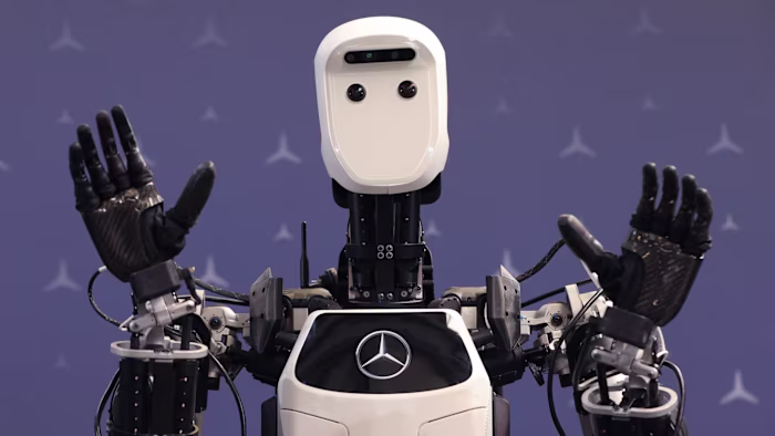 Apollo humanoid robot prototype by Apptronik Inc. raises its hands in front of a backdrop with Mercedes-Benz logos.