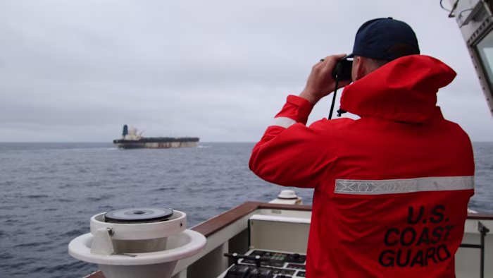 A U.S. Coast Guard member in an orange jacket observes a distant oil tanker through binoculars from a ship’s deck.