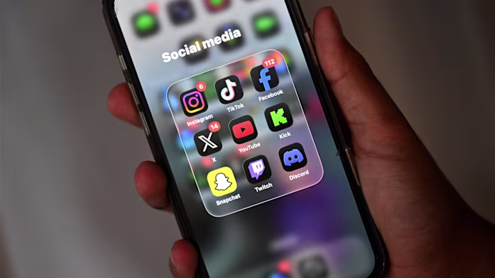 A boy holds a smartphone showing social media apps including Instagram, TikTok, Facebook, X, YouTube, Kick, Snapchat, Twitch, and Discord.