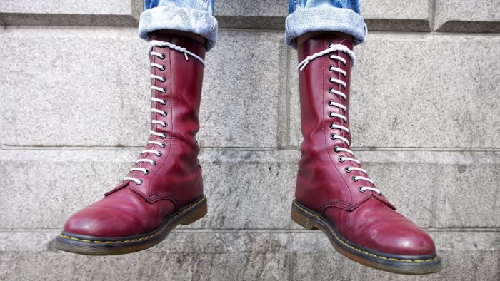 Burgundy Dr Martens boots with white laces worn by someone in cuffed blue jeans, standing against a stone wall.