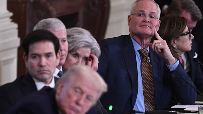 Darren Woods sits at a table with other executives as Donald Trump appears blurred in the foreground.