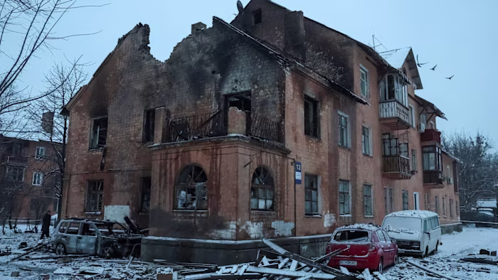 A damaged apartment building with blackened walls and shattered windows, burned vehicles and debris covered in snow in front.